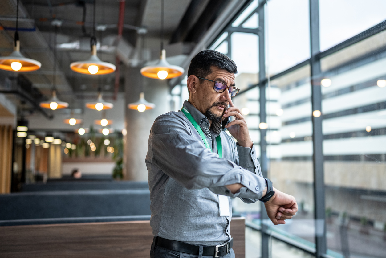 Mature man talking on the phone and checking the time on his wristwatch at office