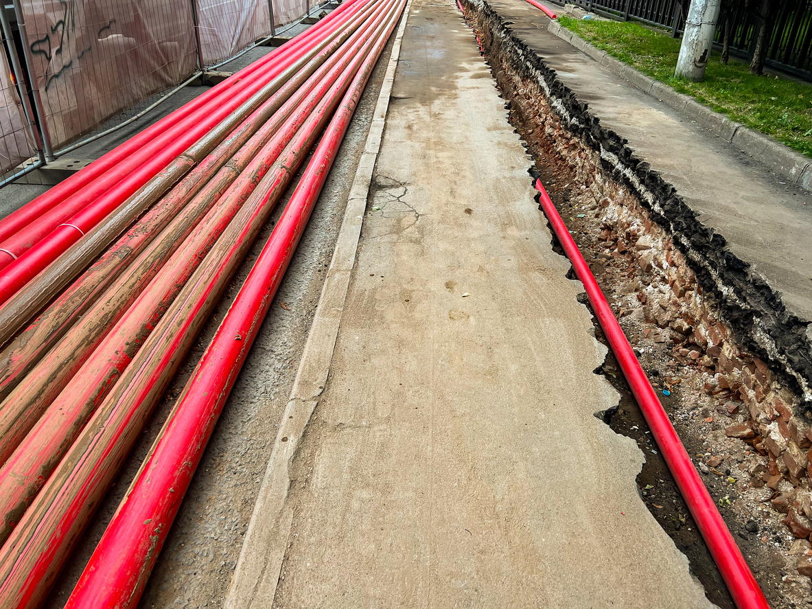 Red plastic pipes stacked on pavement and one pipe in a trench